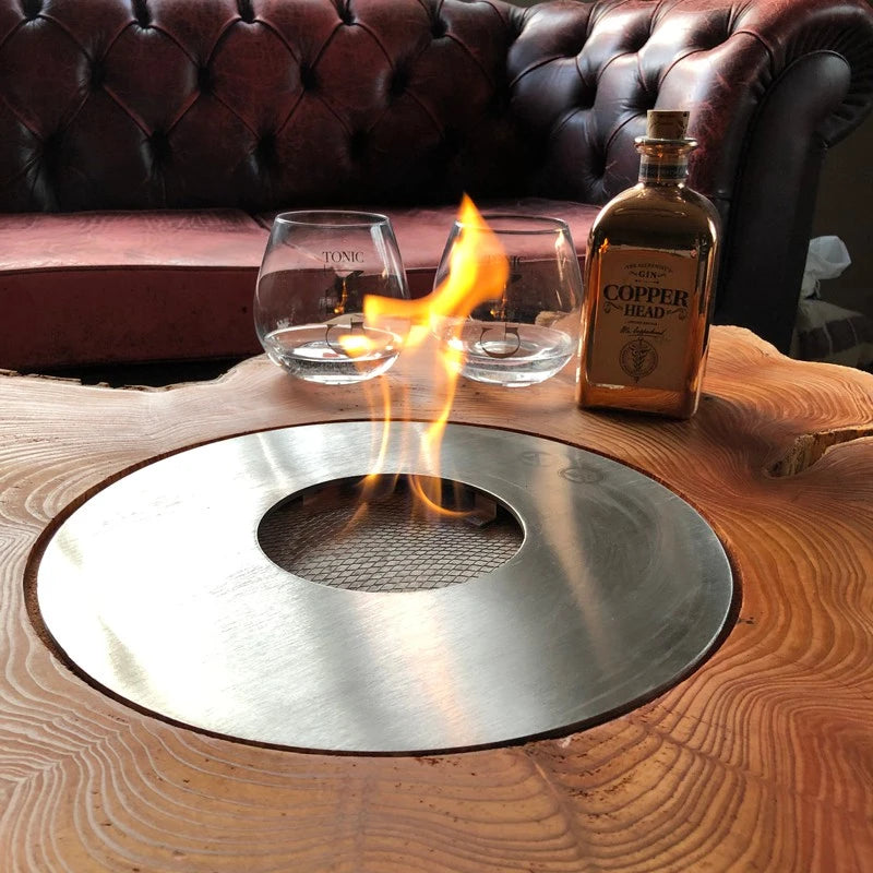 Round 5L outdoor bioethanol burner built into a live-edge wooden table with a lit flame, glasses and a gin bottle, customer photo