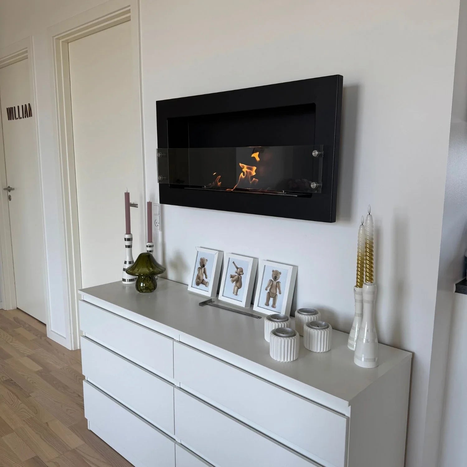 Texas black bioethanol fireplace mounted in a real hallway above a white dresser with framed teddy bear photos and candles, lit with a realistic flame